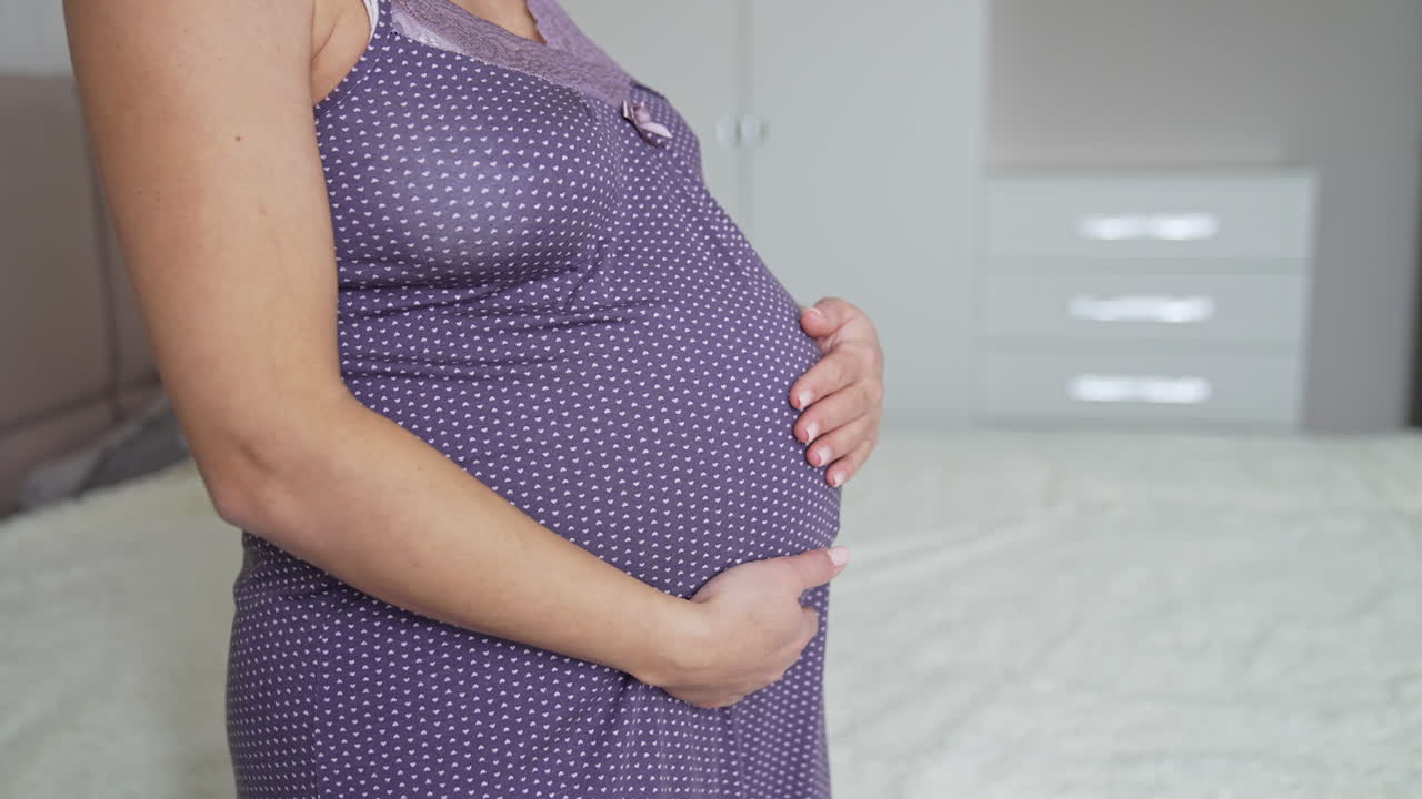 Expecting mommy wearing a nightgown is standing in the bedroom. Woman stroking tenderly her large belly with two hands. Close up.