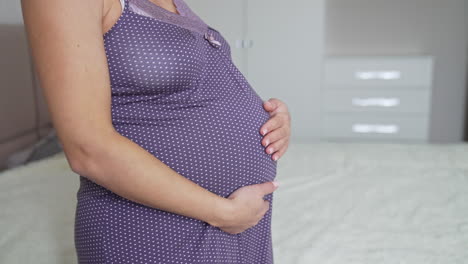 expecting mommy wearing a nightgown is standing in the bedroom. woman stroking tenderly her large belly with two hands. close up.