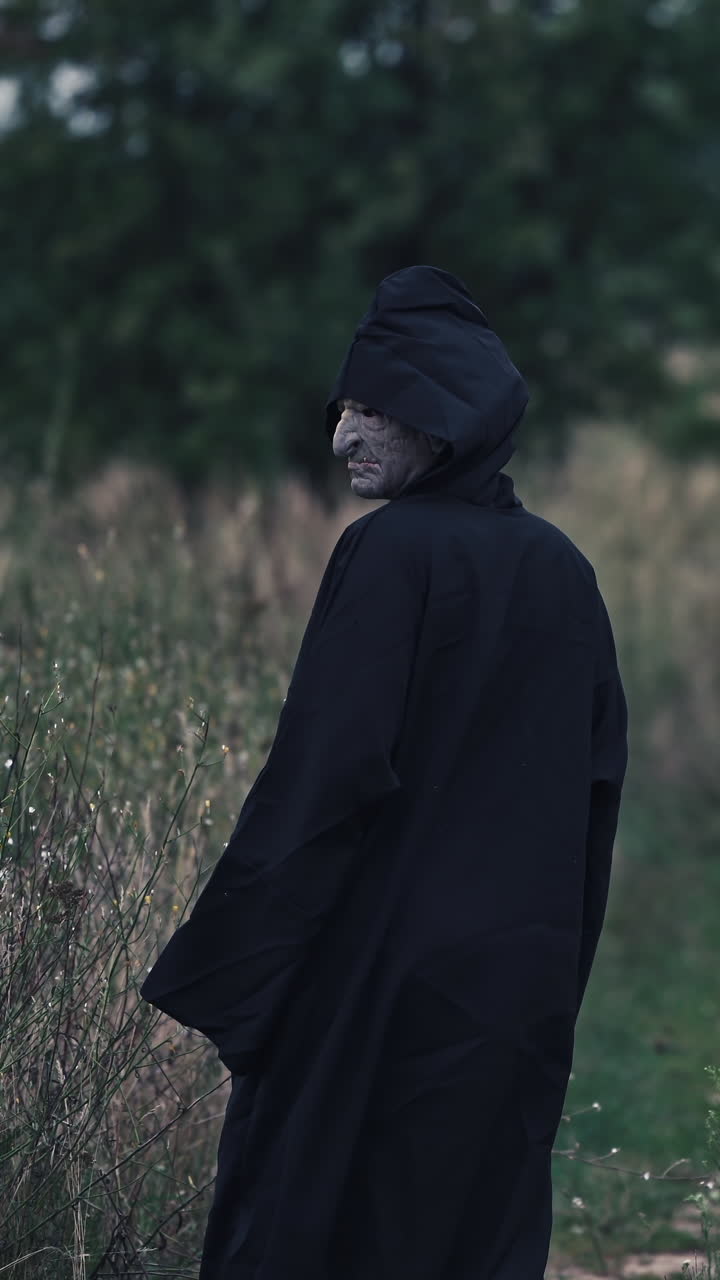 Backside portrait of death in nature. Dark figure in black costume out stretches its arms and goes away in the field in the evening. Scary nightmare ghost outdoors. Vertical video