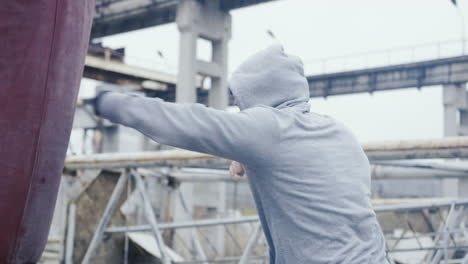 side view of caucasian man in sportswear hitting a punching bag outdoors an abandoned factory on a cloudy morning