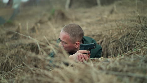 a close view of a man lying on the ground in a dry, grassy area, holding a handgun while aiming with it and looking intensely around