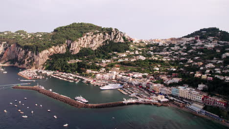 an extreme wide drone shot circling marina grande on the island of capri in campania, italy, showing boats and a ferry in the port and tourists strolling the coast near colorful pastel buildings