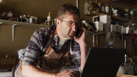 focused caucasian male knife maker in workshop using laptop and smartphone