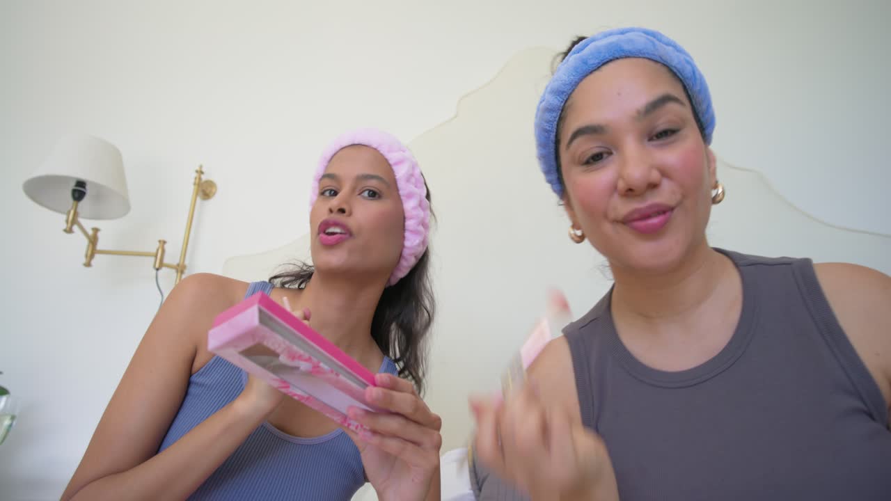 Un duo femminile di diverse origini tiene una palette rosa sul letto, realizza un tutorial di trucco e applica il fard con i pennelli.