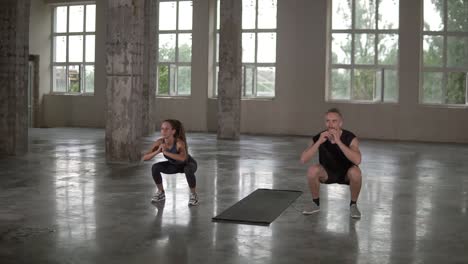 Young-man-and-woman-with-dreadlocks-are-workouting-at-empty-studio,-doing-synchronous-squats-together,-strong-and-healthy,-maintain-their-active-lifestyle,-slow-motion