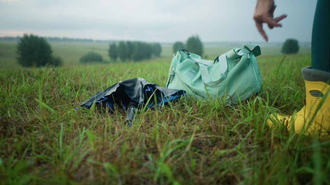 Close-up of partial view of someone in yellow boots dropping an umbrella next to a green bag on a grassy field, trees are visible in the far distance under a cloudy sky