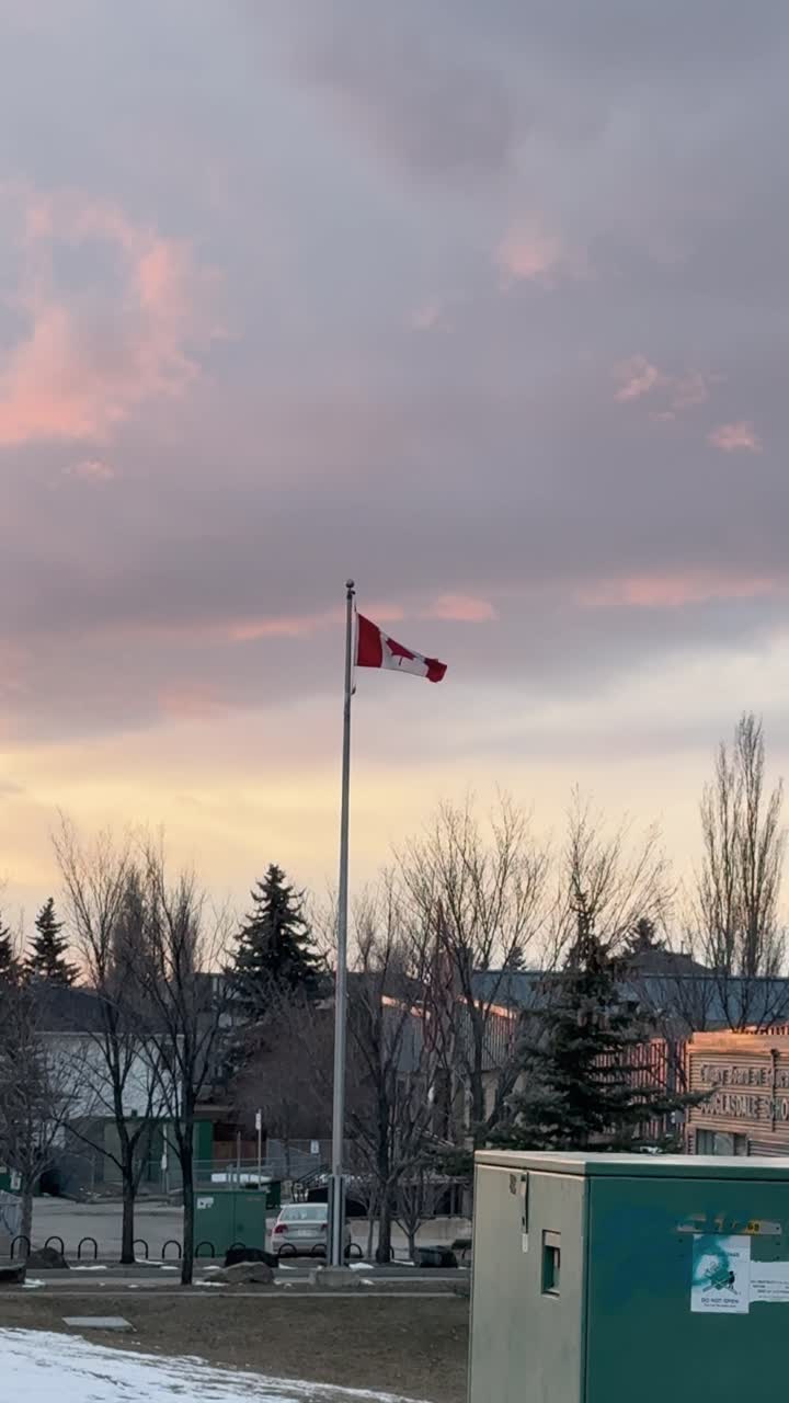 Una bandiera canadese sventola in cima a un alto palo contro un cielo al tramonto dai colori pastello. Alberi spogli ed edifici vicini incorniciano la scena in un tranquillo paesaggio urbano serale.