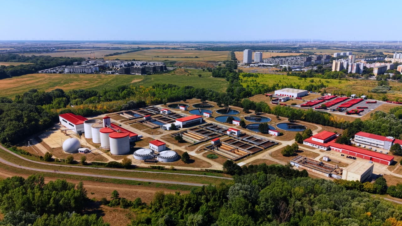 Wastewater treatment facility beside fields and housing. An elevated view shows tanks and basins surrounded by trees, with apartment blocks in the distance.