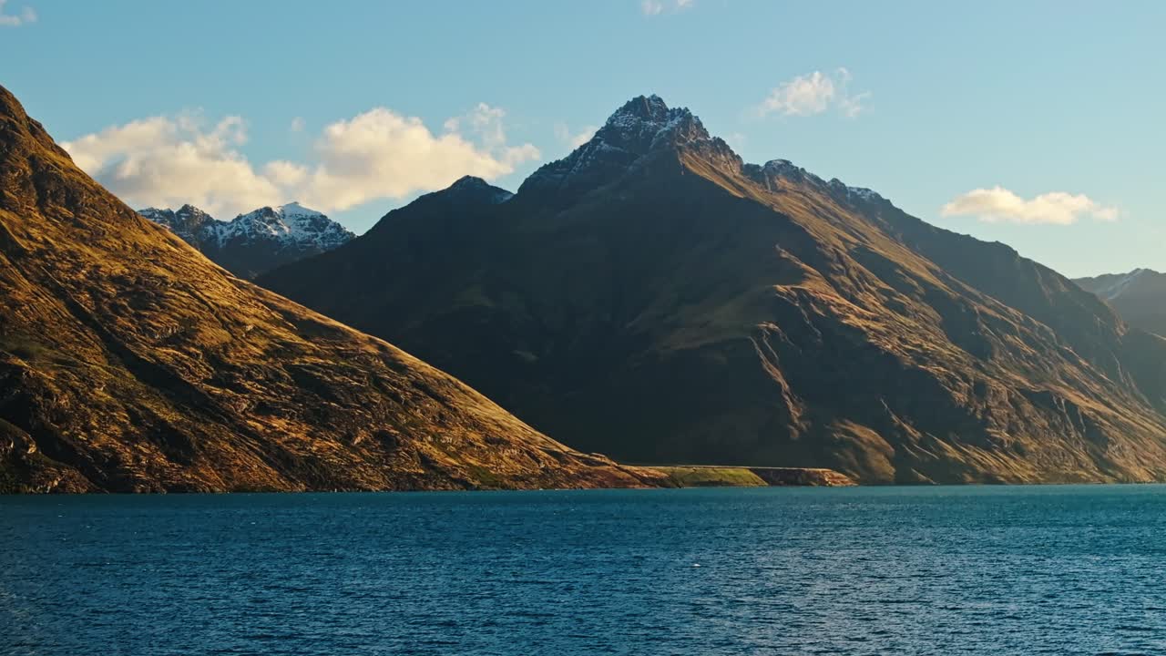 Aerial pan across red brown hills sloping into Lake Wakatipu with snow covered peaks