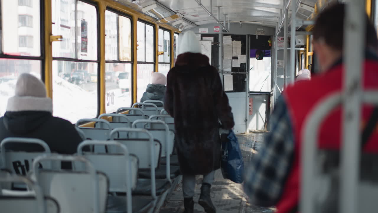 man hands ticket to woman near door on moving winter train, commuters bundled in seats, corridor perspective, soft daylight through windows, urban transit moment before stop
