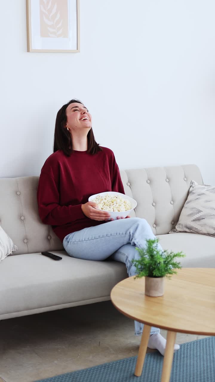 Giovane donna felice che ride mentre guarda la TV e mangia popcorn sul divano
