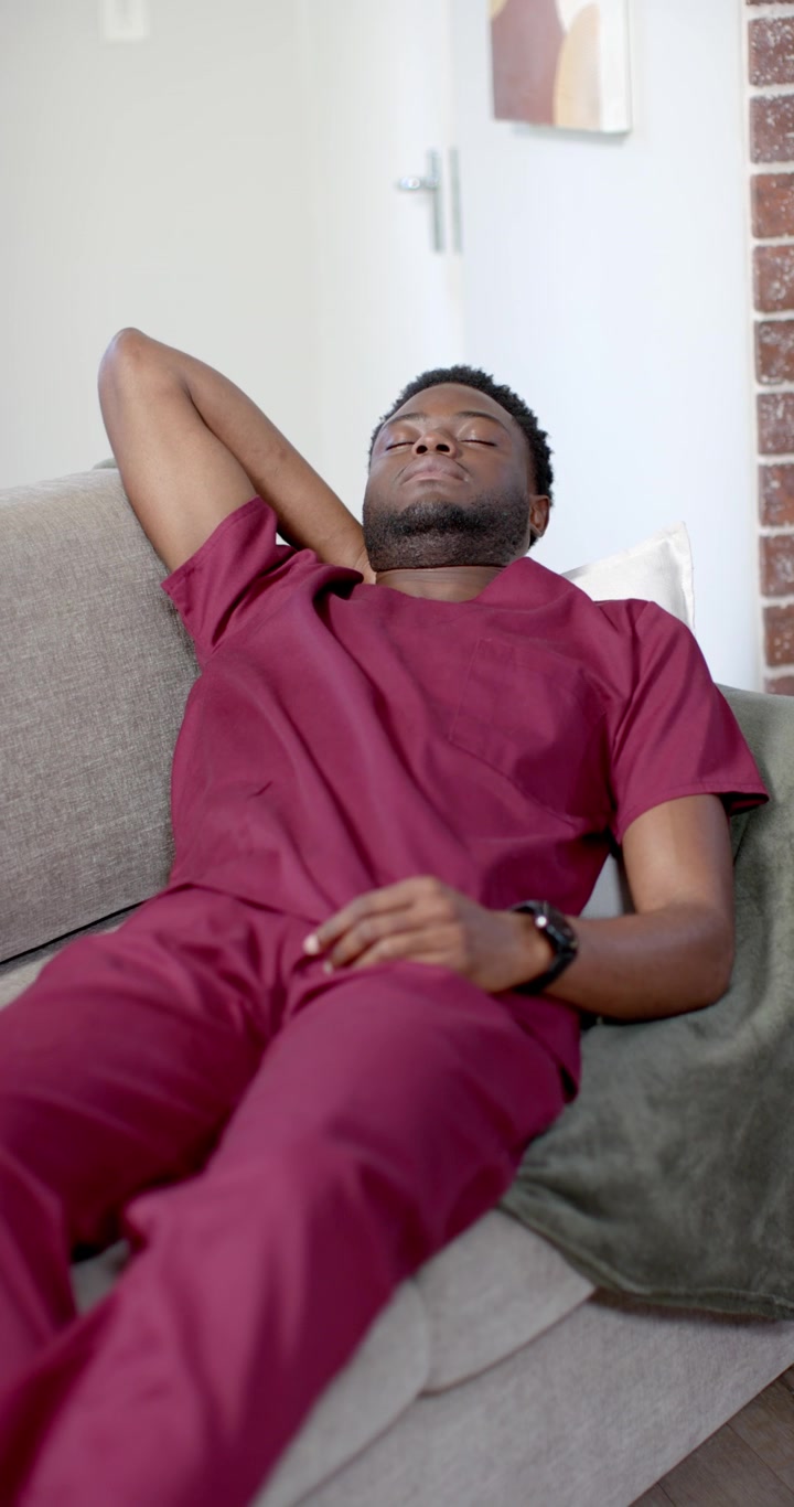 Vertical video: African man lying at home on sofa breathing, settling, napping in scrubs with watch