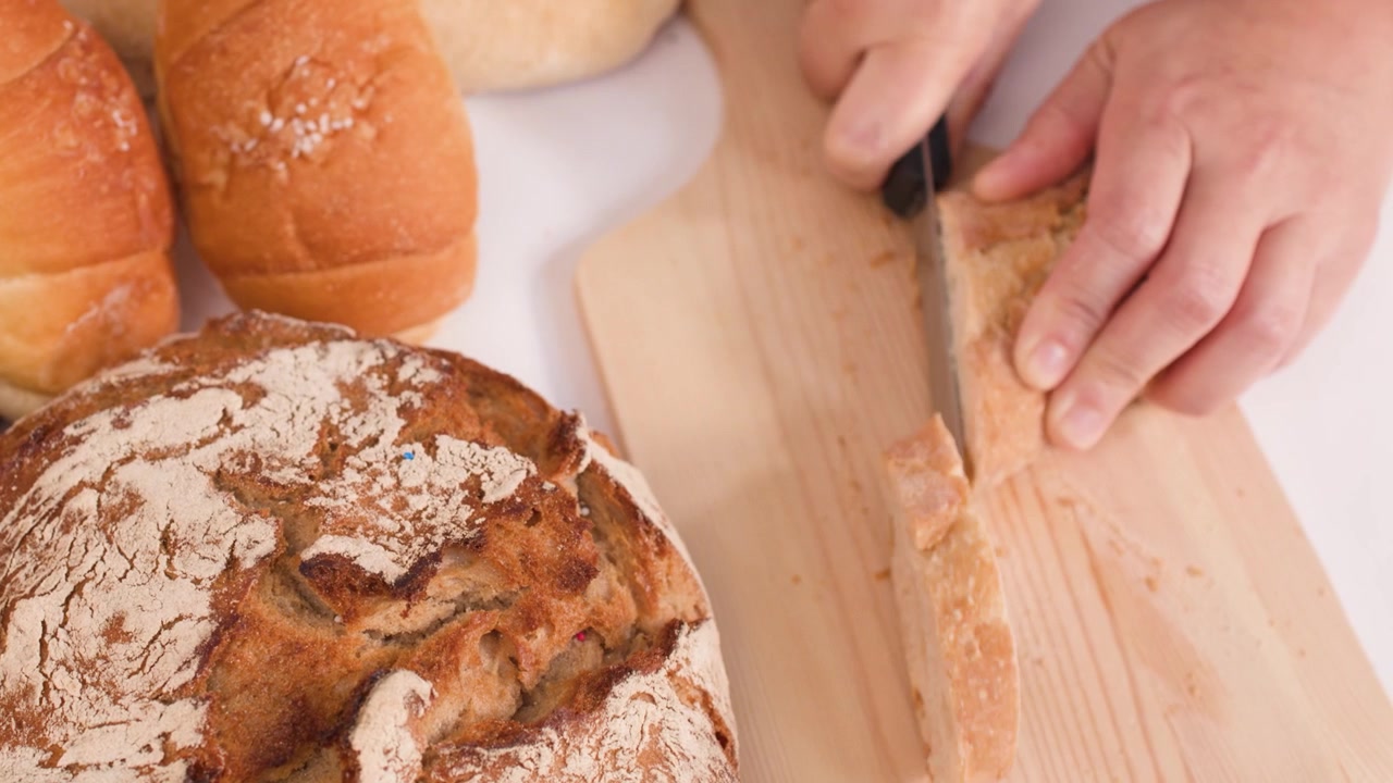 Close-up overhead shot of hands slicing crusty baguette beside sourdough and artisan rolls.
