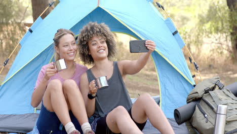 Dos-Mujeres-Sentadas-Dentro-De-Una-Tienda-De-Campaña,-Una-Sosteniendo-Una-Taza-Y-La-Otra-Tomándose-Una-Selfie.