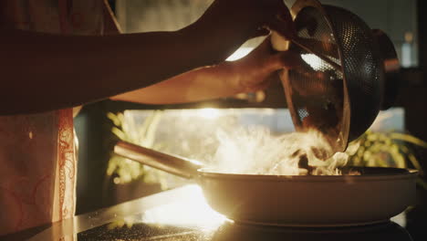 a woman pours wild mushrooms into a hot frying pan. the sun from the window beautifully illuminates the stove and pan