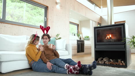 christmas time, couple enjoying hot cocoa by fireplace wearing festive reindeer antlers at home