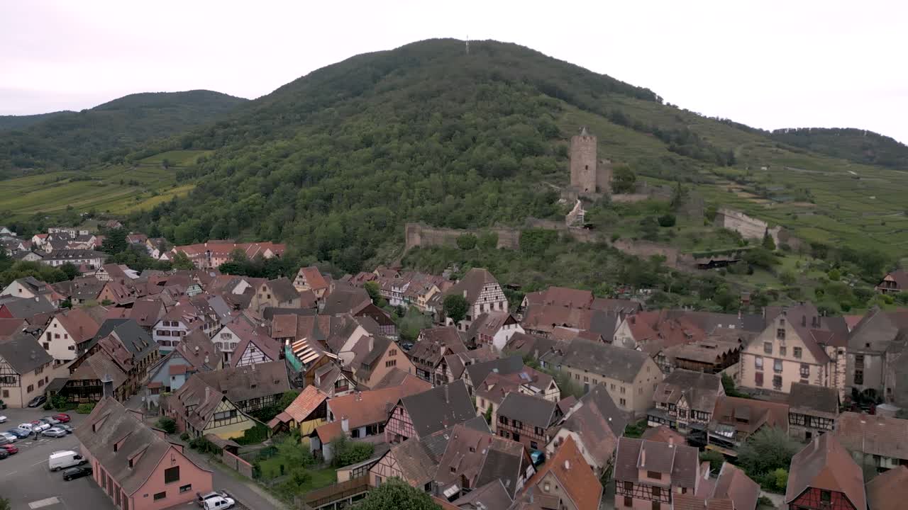 Aerial drone shot of a traditional Alsatian village nestled at the foot of forested hills, featuring half-timbered houses, red rooftops, and the ruins of a medieval stone castle on the hill