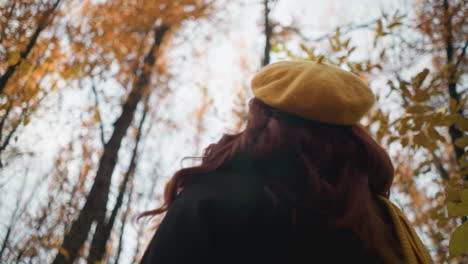 elegant lady in yellow beret and muffler holds vibrant autumn leaf over face, shielding from sunlight while gazing up at tall trees with golden foliage