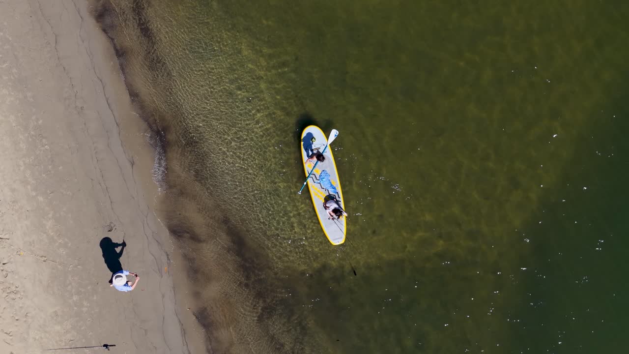 Kids fish and paddleboard near sandy shore, aerial drone view, bright daylight, calm atmosphere