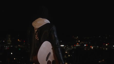 younger musician with guitar stands in front of city lights in the night