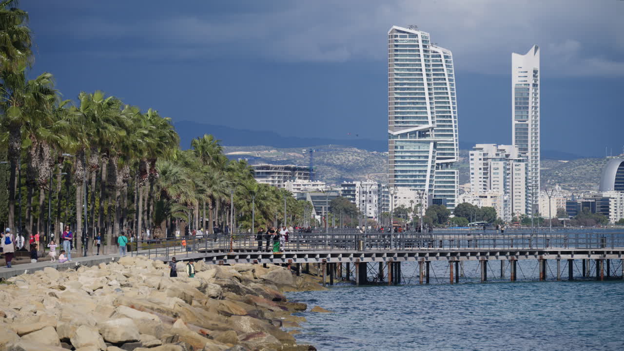 Limassol, Cipro - 26 febbraio 2026: Pontile di legno che si estende nel Mar Mediterraneo a Limassol con persone che camminano e moderni edifici a molti piani sullo sfondo.