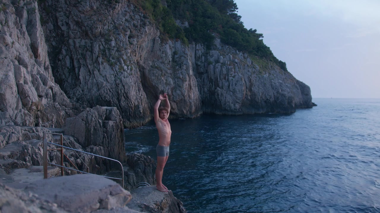 Italian man stands on rocks at blue hour, preparing to jump into the ocean at Gradola Beach, Capri Island, Italy