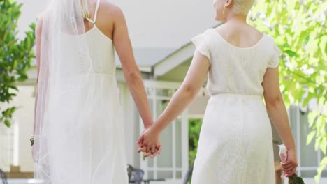 Happy-diverse-female-couple-holding-hands-and-bunch-of-flowers-in-garden