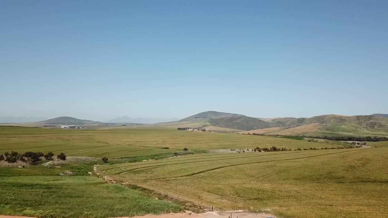 Toma de dron volando sobre tierras de cultivo con colinas al fondo y un coche azul conduciendo por un camino de tierra en primer plano