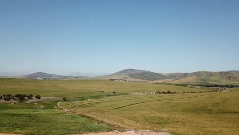 disparo de drones volando sobre tierras de cultivo con colinas en el fondo y conducción de automóviles azules en un camino de tierra en primer plano