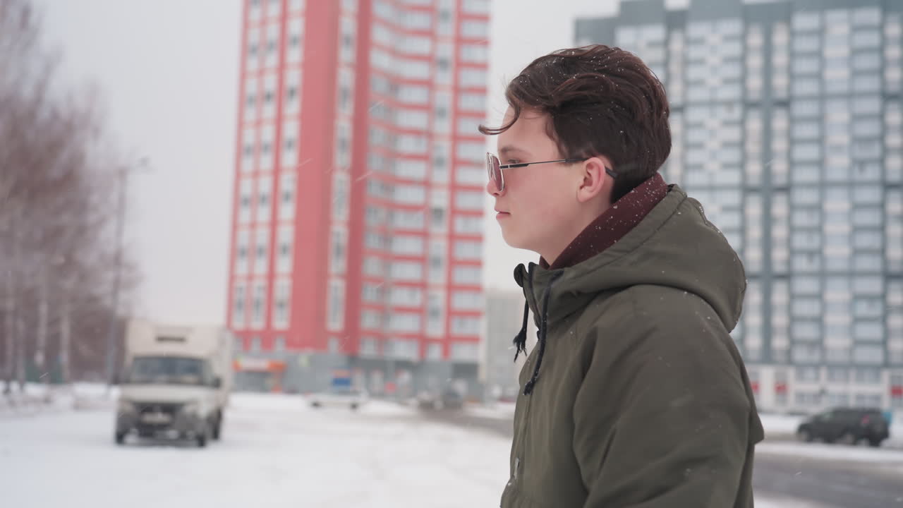 Side view of young man in winter jacket walking toward parked car and entering vehicle during snowfall with moving cars and truck visible near tall buildings in urban snowy parking lot