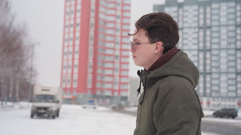 side view of young man in winter jacket walking toward parked car and entering vehicle during snowfall with moving cars and truck visible near tall buildings in urban snowy parking lot