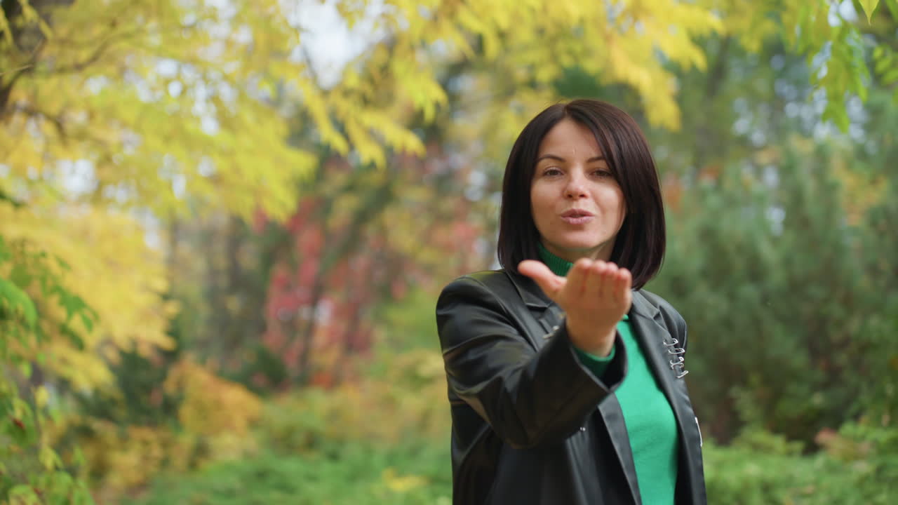 Mujer con chaqueta de cuero caminando entre el follaje otoñal, con un jersey verde asomando, girándose para lanzar un beso y estirando la mano, hojas doradas, sendero arbolado, estilo rock casual, sonrisa cálida y mirada directa