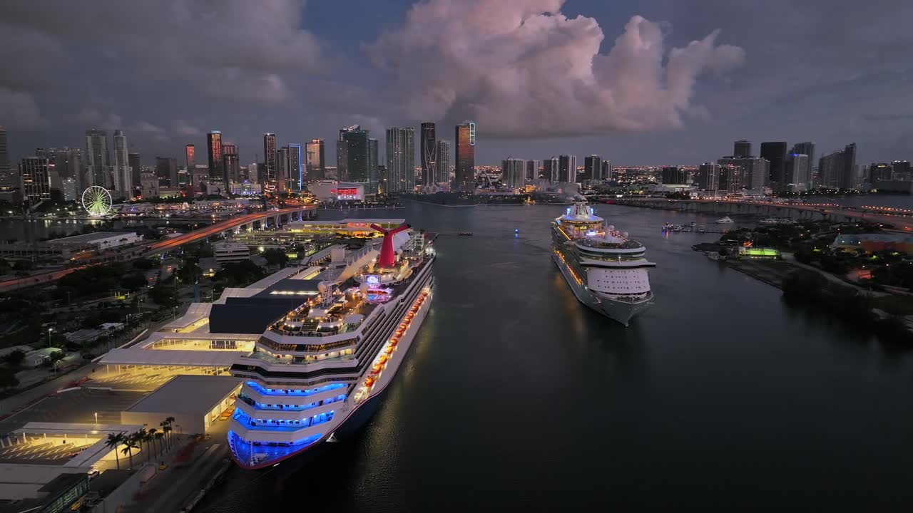 Cruise ships docked at Port of Miami with the brightly lit downtown Miami skyline in the background. The shot overlooks the port and Biscayne Bay as night falls. Aerial.