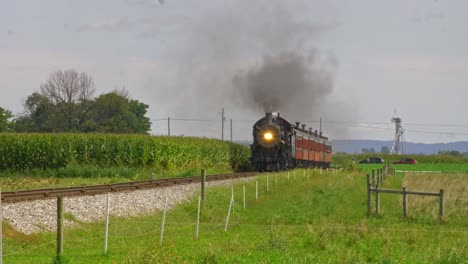 una vista de un tren de pasajeros de vapor antiguo que sopla humo y vapor que viaja a través de campos de maíz fértiles en un día soleado de verano