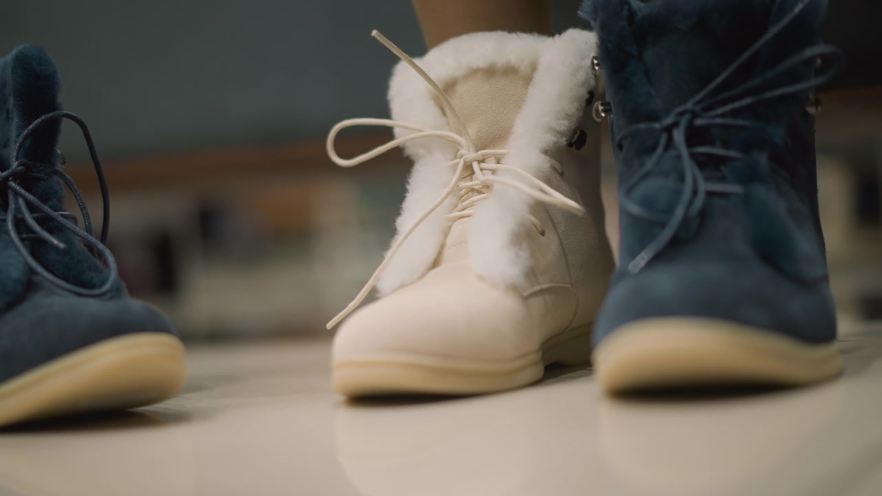 Close-up dua sepatu bot musim dingin, satu biru dan satu putih, dengan hiasan bulu, diletakkan berdampingan di permukaan halus. Pemandangan menunjukkan detail tekstur termasuk tali yang diikat pita pada kedua sepatu.