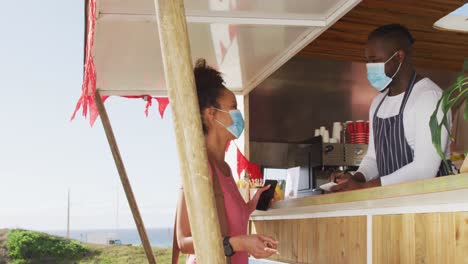 African-american-male-food-truck-owner-taking-order-from-female-customer,-both-in-face-masks