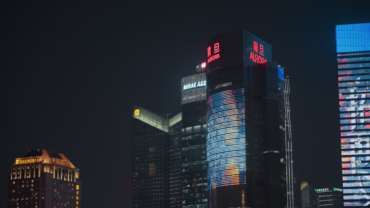 Grattacielo con facciata a LED di notte, skyline cittadino scintillante con display pixel animato sulla torre di vetro, toni vibranti di blu e rosso, movimento pubblicitario commerciale, atmosfera di vita notturna urbana, aerea