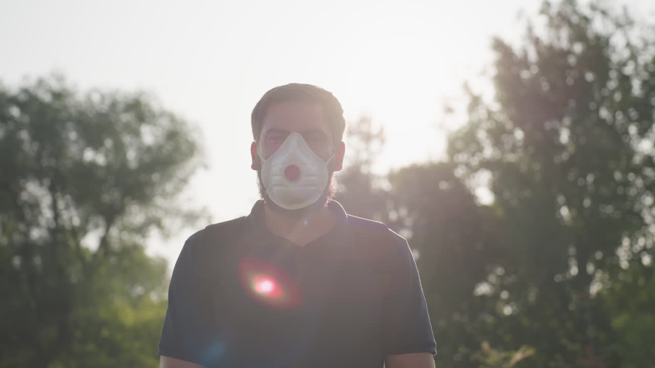 man gardener wearing mask facing camera under bright sunlight creating lens flare while performing outdoor fumigation in field with green plants and trees background