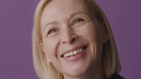 Close-Up-View-Of-Blonde-Mature-Woman-With-Green-Eyes-Smiling-And-Looking-At-Camera-On-Purple-Background