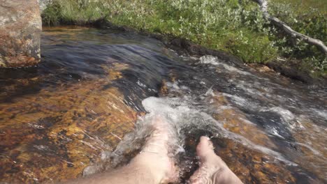 male hiker dipping feet in river stream running water, point of view pov