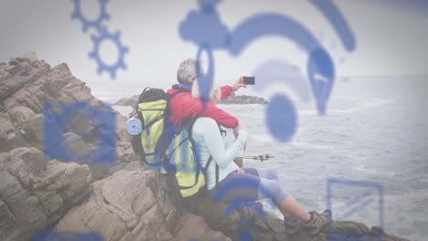 animation of digital online icons over senior couple by seaside taking selfie with smartphone