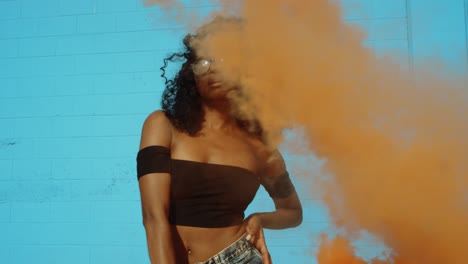 beautiful young woman holding colorful smoke grenade dancing outside against blue wall