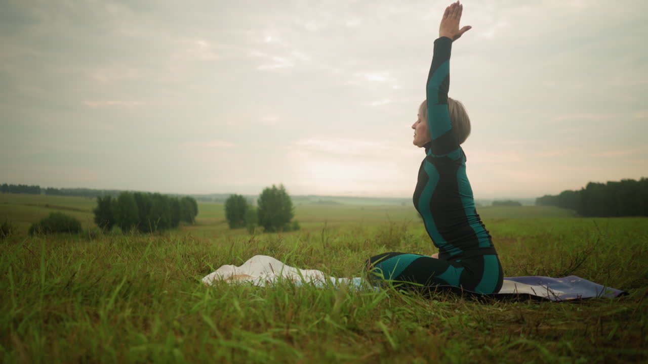 Woman in green and black suit seated on yoga mat with legs crossed and hands joined, stretching upward in grassy outdoor field, she bends forward with focus, practicing yoga under cloudy sky