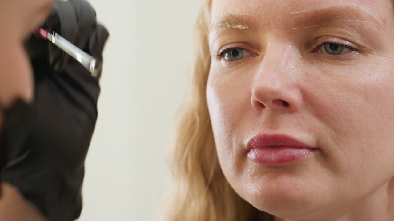 Close-up view of client with focused expression as technician applies cream to eyebrow using precision brush, technician face partially blurred in foreground for artistic depth effect