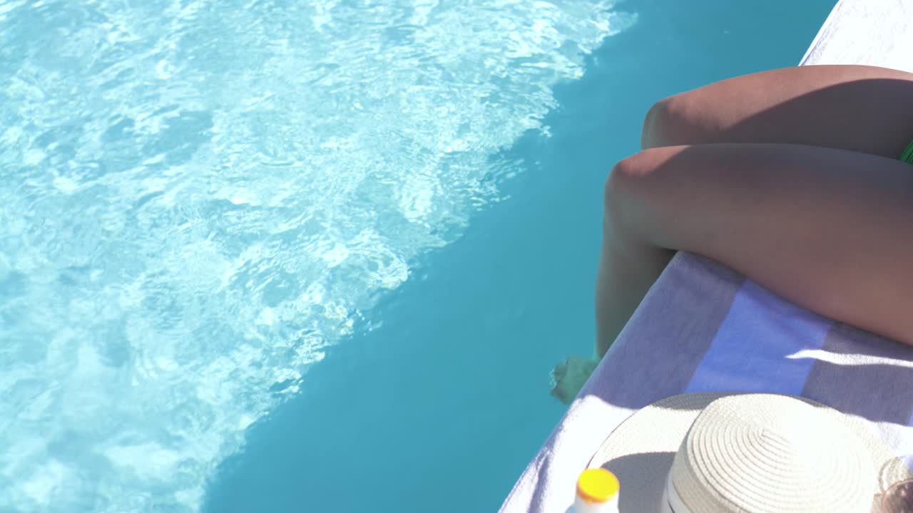Woman sitting towel at pool edge dipping feet, cooling from sun with sunscreen nearby, copy space
