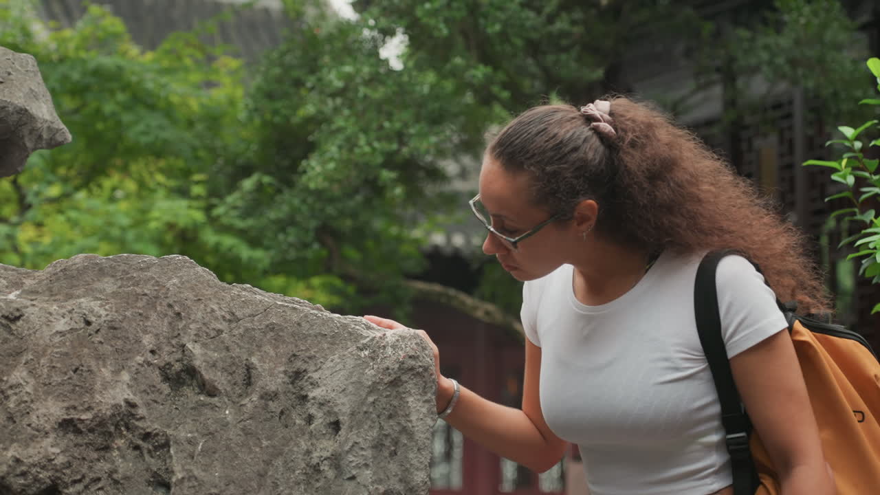 Donna multietnica ispeziona da vicino una pietra, con lo sguardo attento di una ricercatrice mentre tocca il lichene e ne studia la texture superficiale accanto agli alberi del cortile; occhiali e taccuino suggeriscono un lavoro sul campo educativo e un'atmosfera tranquilla.