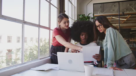 Three-millennial-women-working-together--at-a-desk-in-a-creative-office,-front-view,-panning-shot