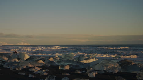 a cold, icy shoreline with floating ice chunks reflecting the last light of sunset, surfer walks out with waves crashing behind