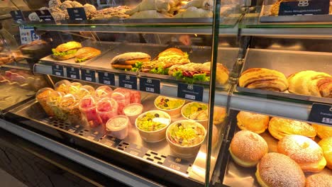 camera pans across a well-lit bakery display case in berlin, showcasing assorted fresh pastries, bread rolls, sandwiches, and desserts under warm lighting