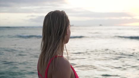 beautiful girl in red swimsuit walks out into ocean during sunset and lightly runs her harnds through the water in hawaii